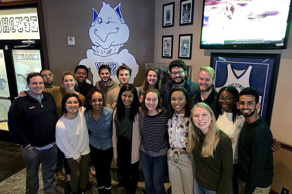 A group of students pose for a photo with a Billiken on the wall behind them.