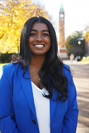 A headshot photo of Reueline Arulanandam with a clocktower and trees in the background.